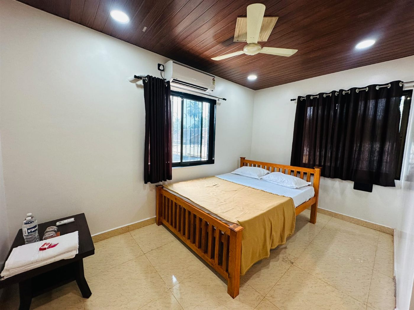 Supari room with wooden double bed, AC, two curtained windows and a side table with towels and drinking water